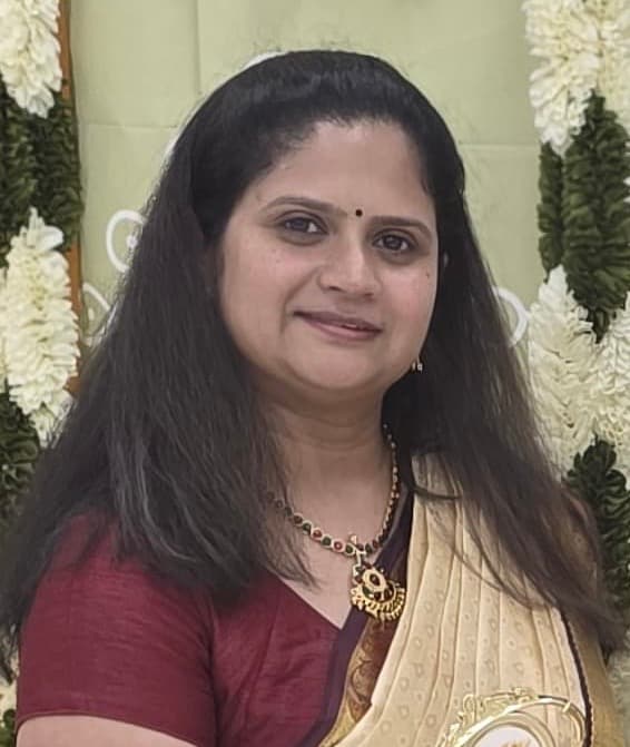 Smiling woman wearing a cream and maroon sari, gold necklace, and bindi against floral garlands.