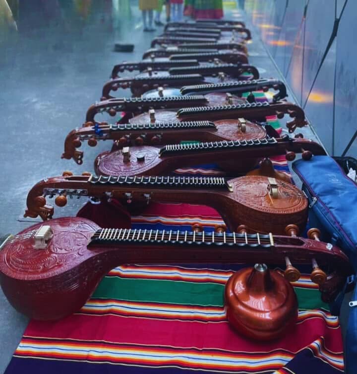 Ornate wooden Saraswati veenas arranged in a long row on a vibrant, striped outdoor rug.