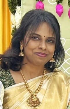 Smiling woman in a gold saree and ornate jewelry standing before festive pink floral decorations.