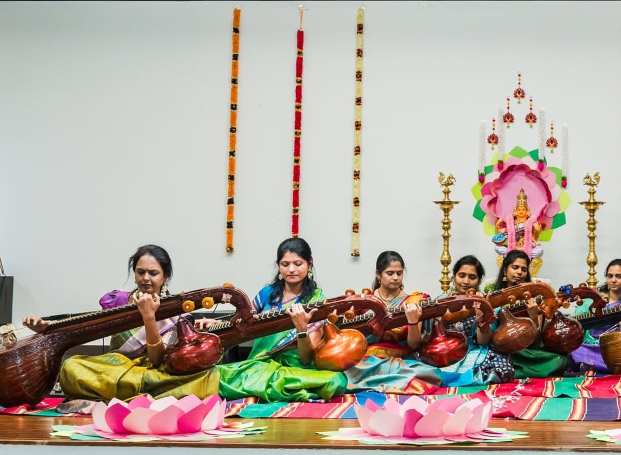 Women in colorful saris play the Saraswati Veena seated on a decorated performance stage.