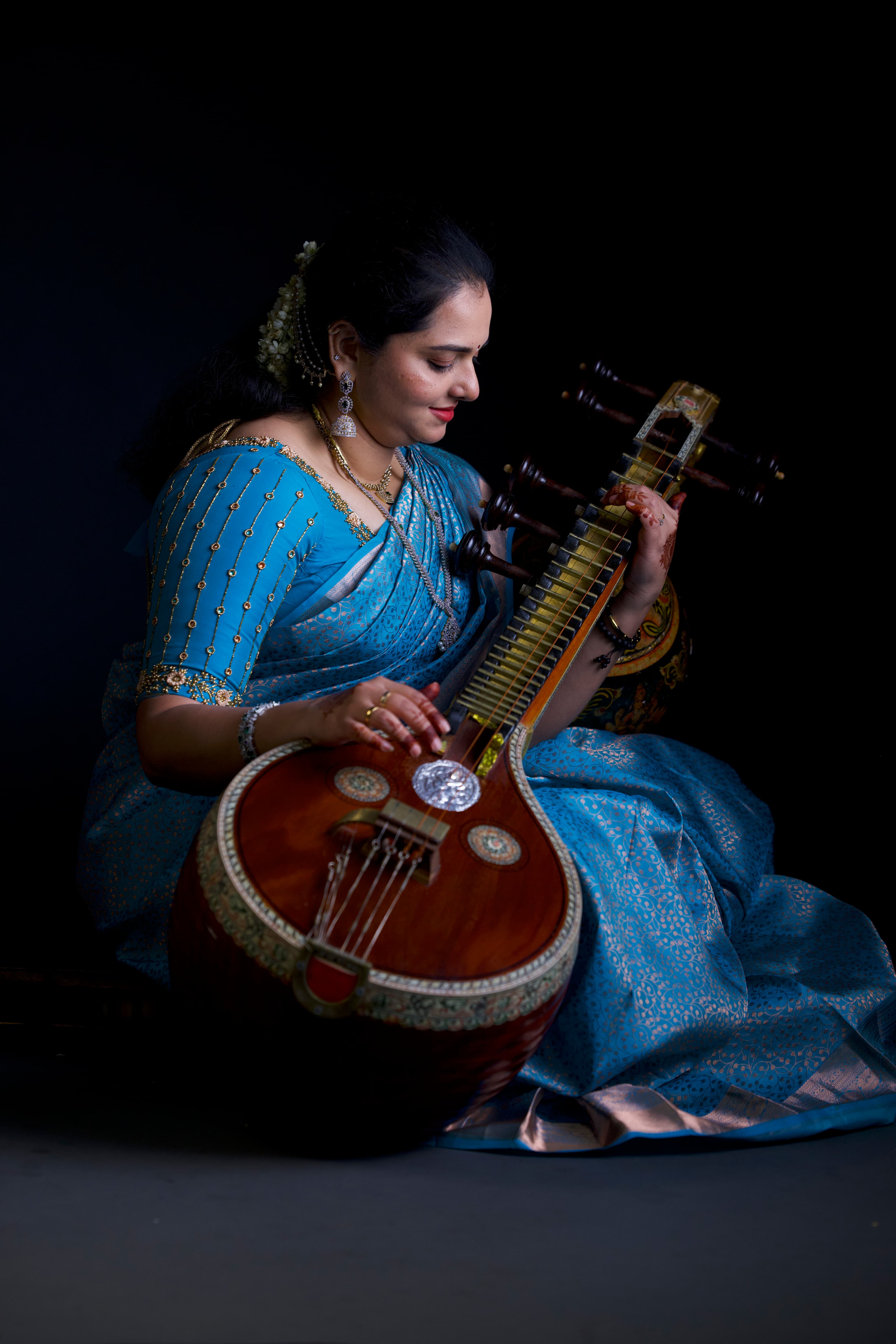 A woman in an ornate blue saree playing a Saraswati veena against a dark background.