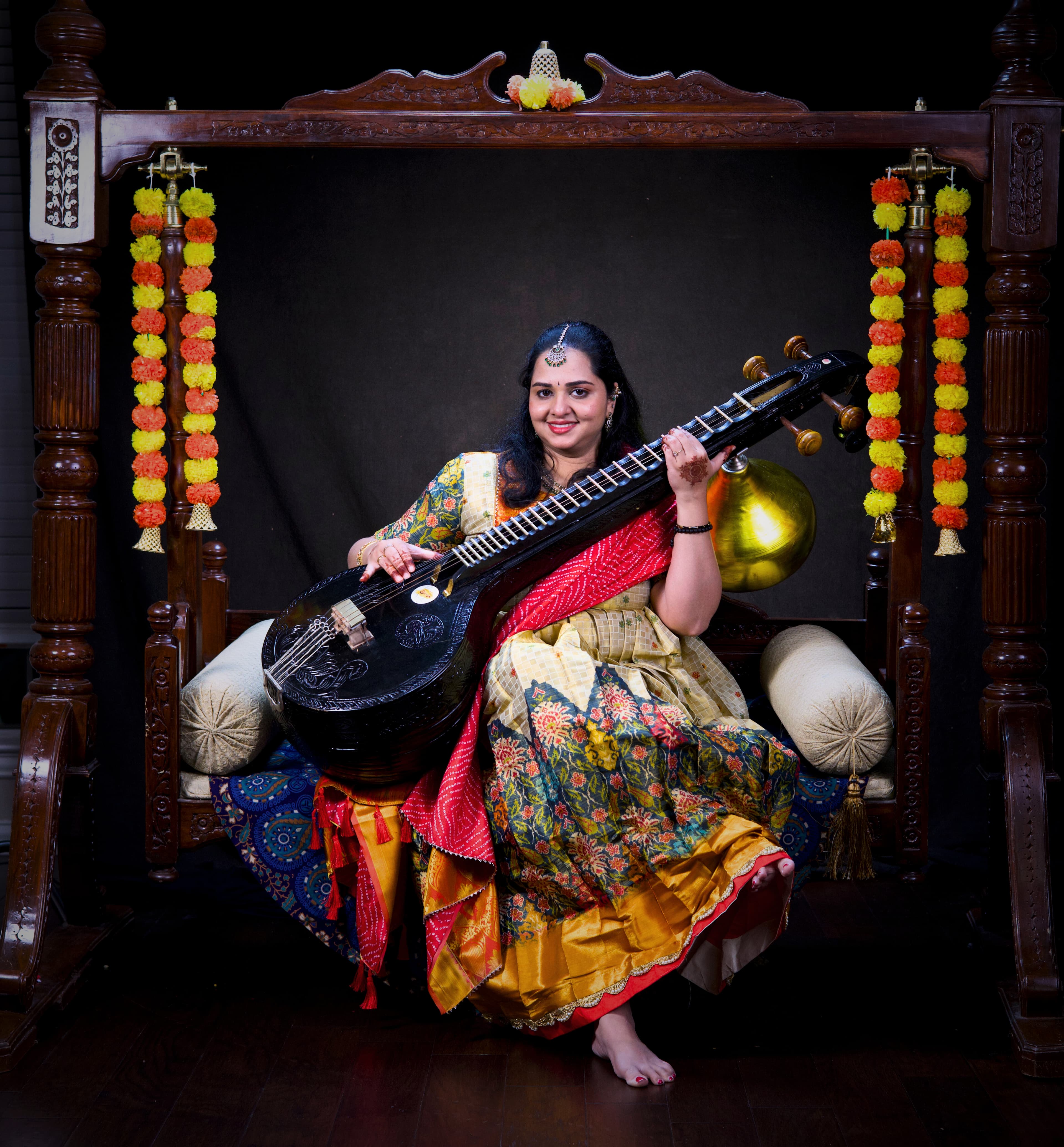 Smiling woman in colorful traditional attire posing with a Saraswati Veena on a wooden swing.