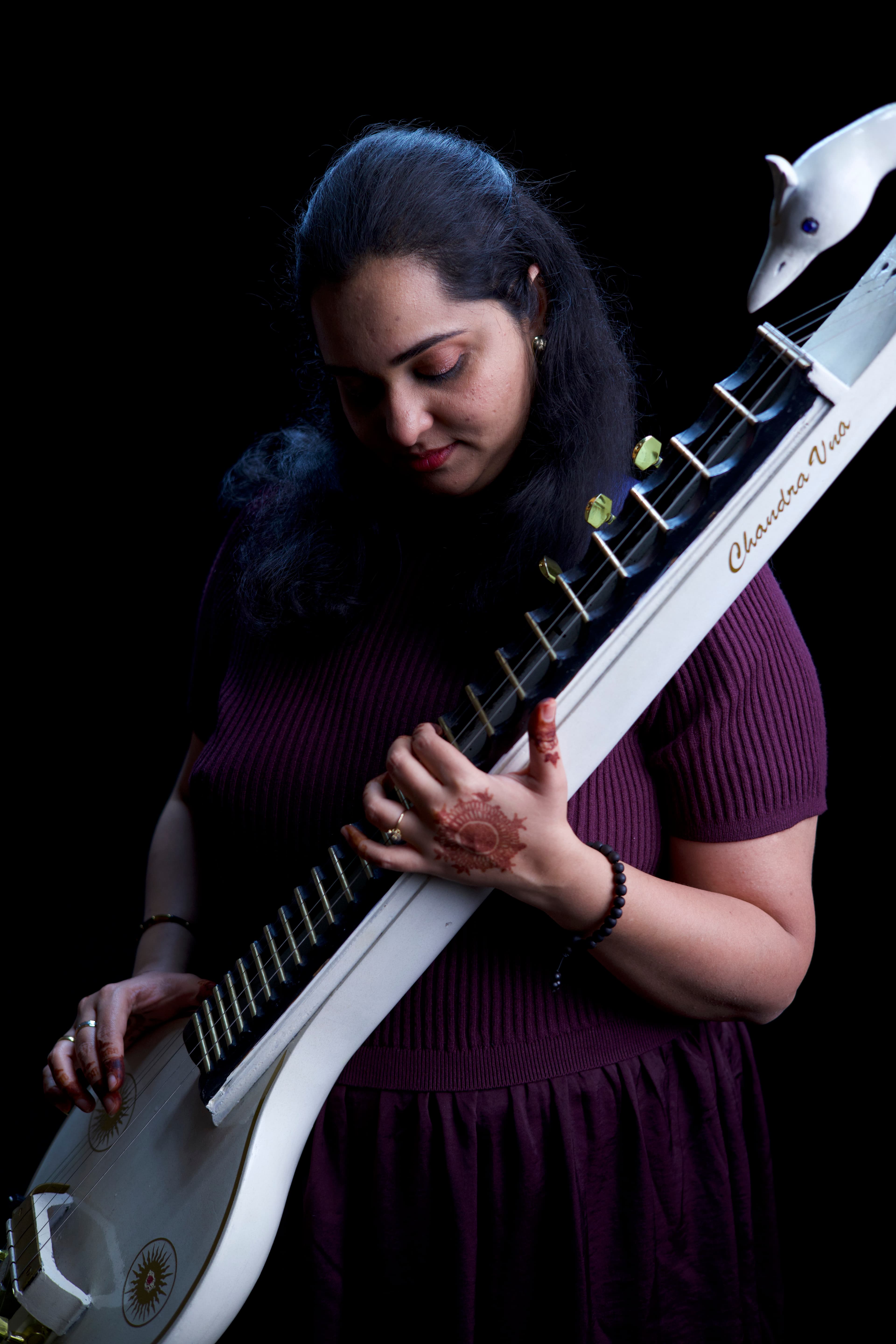 Woman with henna-decorated hands playing a white Chandra Vina against a stark black background.