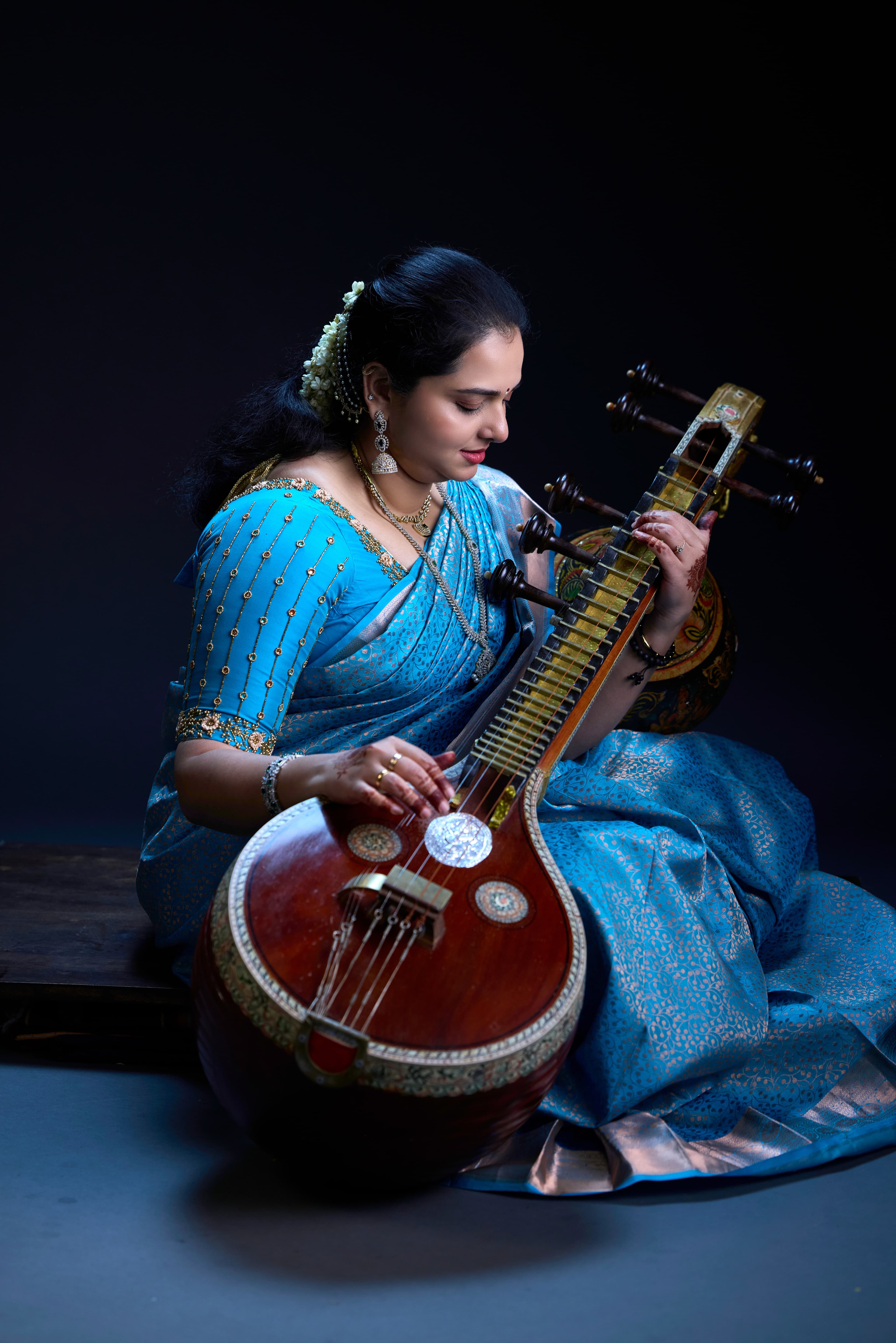 Seated woman in an ornate blue saree playing a traditional Indian Saraswati Veena instrument.
