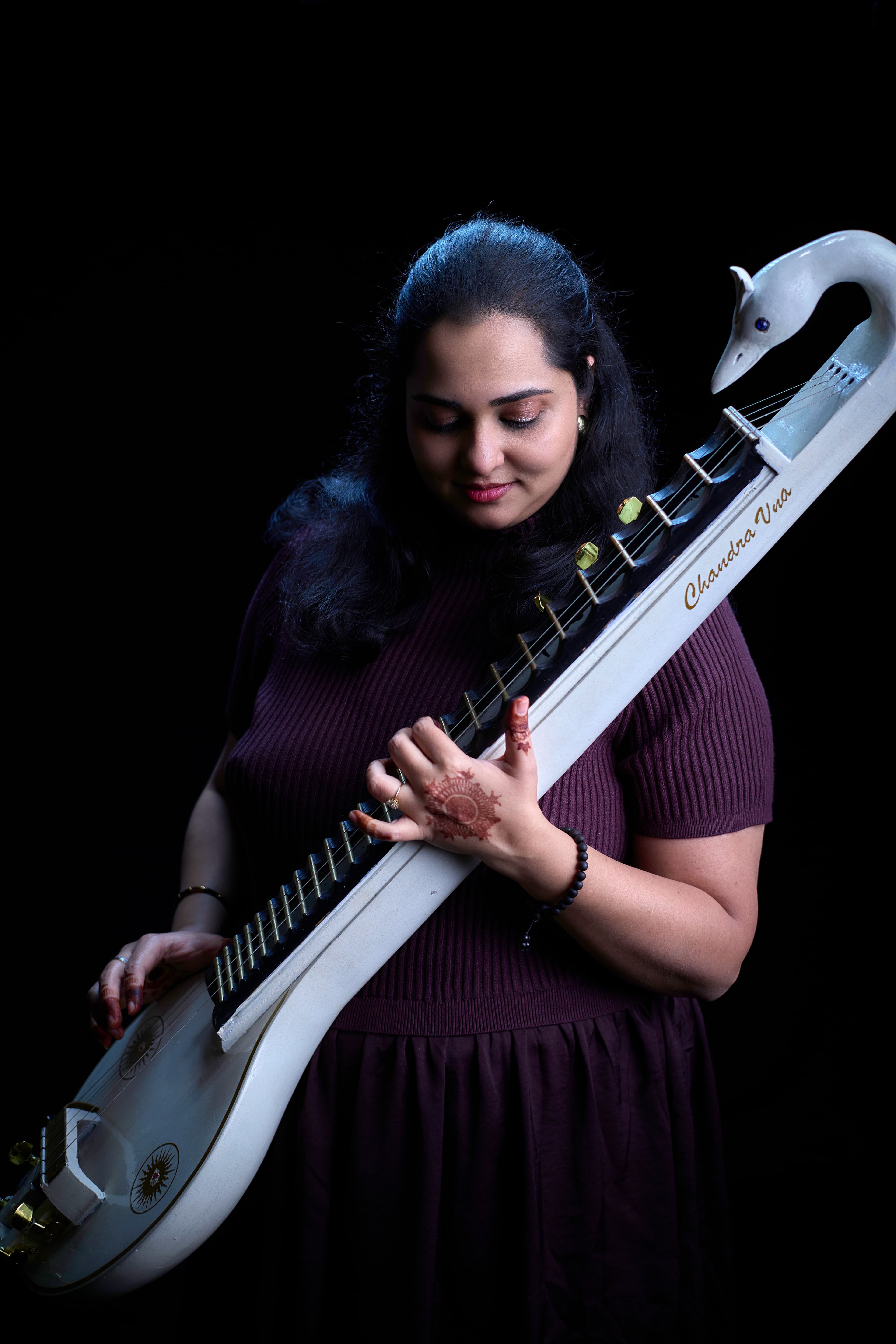 Woman with hennaed hands playing a white swan-headed Chandra Veena against a black background.