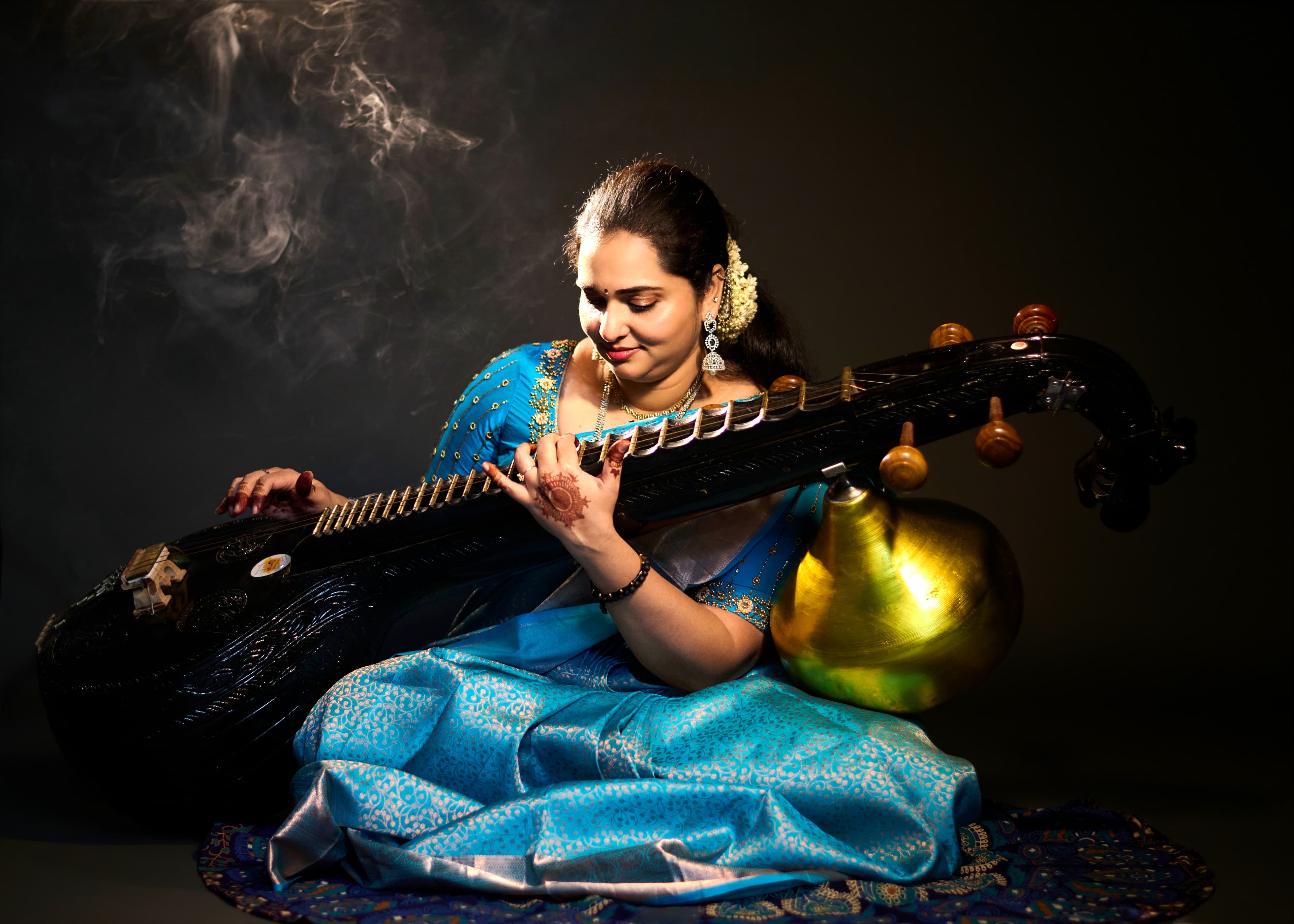Woman in a blue sari playing a Saraswati veena against a dark, smoky background.