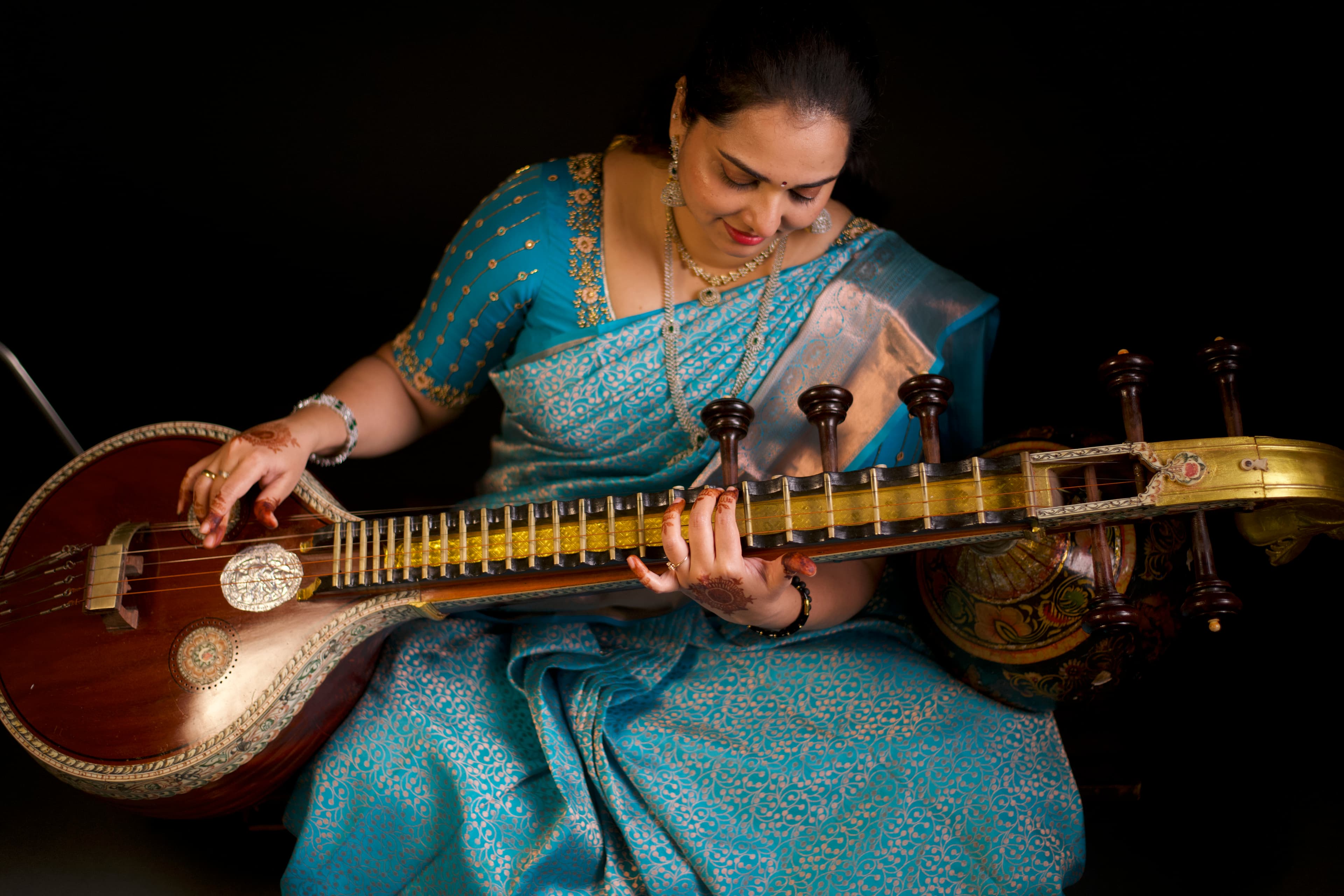 Woman in an ornate blue saree playing a Saraswati Veena against a dark background.