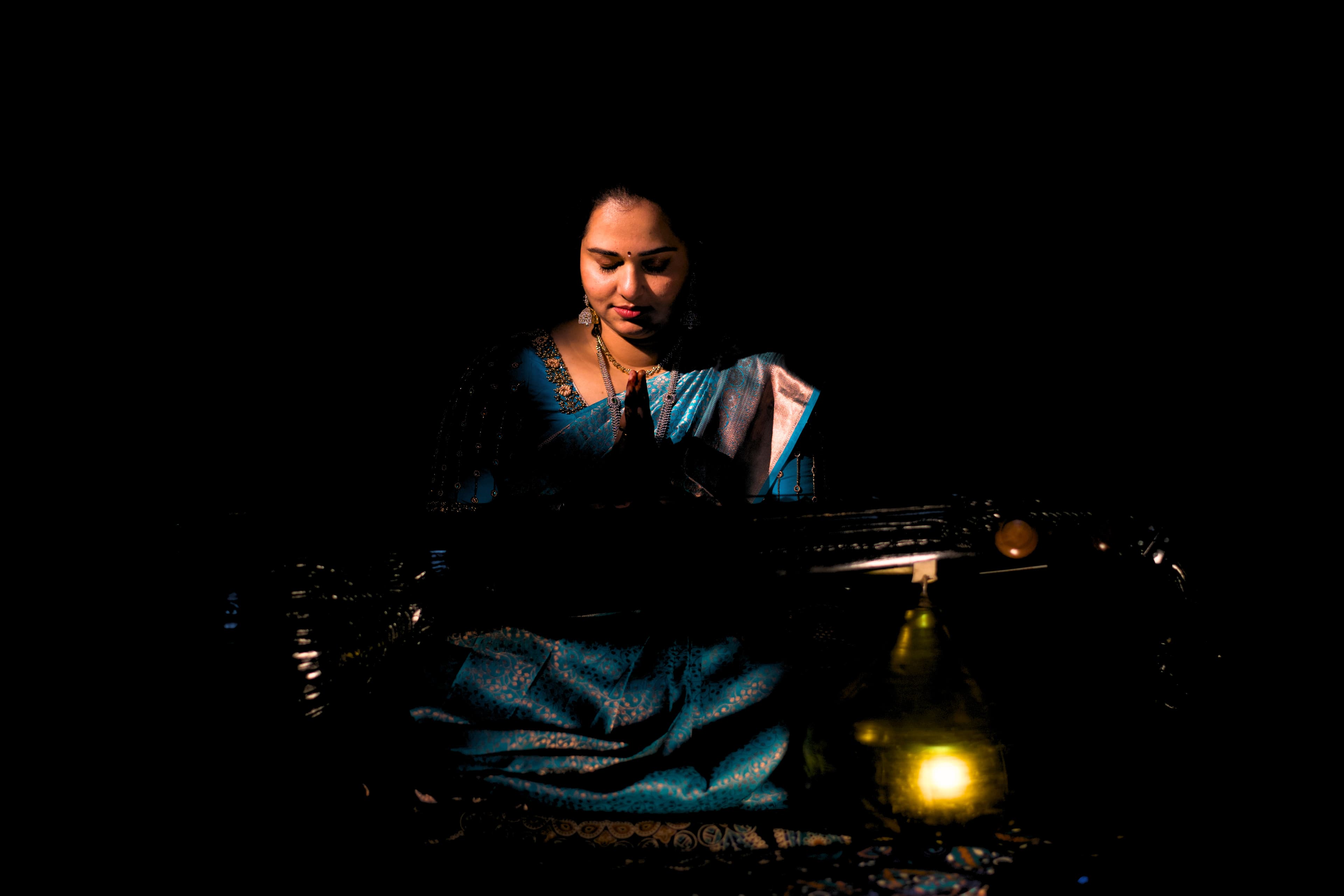 Woman in blue saree with hands in prayer, sitting behind a veena in dramatic lighting.