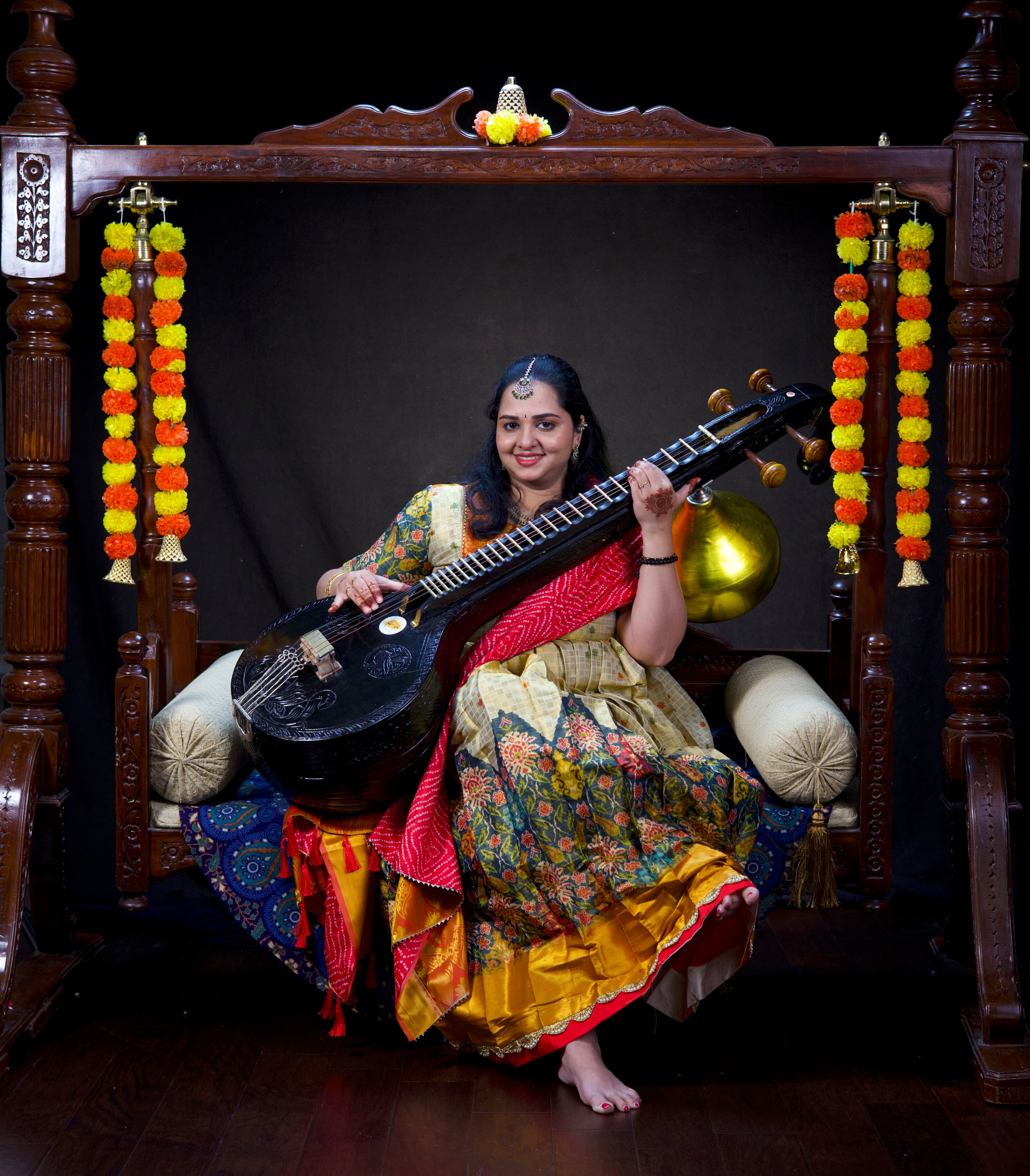Smiling woman in traditional Indian attire sits on a swing holding a Saraswati Veena.
