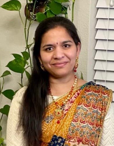Smiling woman with long dark hair wearing a traditional patterned saree and gold jewelry.
