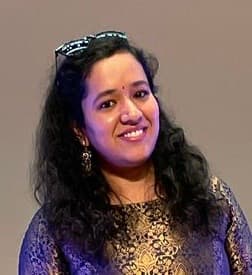 Smiling woman with dark curly hair and glasses on head, wearing a blue patterned top.